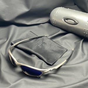 May include: Silver-framed sunglasses with blue lenses and a black carrying case. The sunglasses have a sleek, modern design with the brand name "Oakley" visible on the case and the lenses. The case is silver and has a cylindrical shape.
