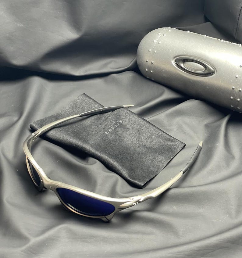May include: Silver-framed sunglasses with blue lenses and a black carrying case. The sunglasses have a sleek, modern design with the brand name "Oakley" visible on the case and the lenses. The case is silver and has a cylindrical shape.