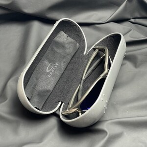 May include: Open gray sunglasses case with a pair of sunglasses inside. The sunglasses have a silver frame and blue lenses. A black microfiber cloth with the Oakley logo is also inside the case.