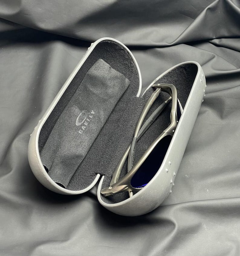 May include: Open gray sunglasses case with a pair of sunglasses inside. The sunglasses have a silver frame and blue lenses. A black microfiber cloth with the Oakley logo is also inside the case.