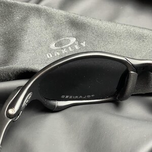 May include: Black sunglasses with a curved frame and dark lenses. The word "POLARIZED" is printed on the lens. A black Oakley carrying case is in the background.