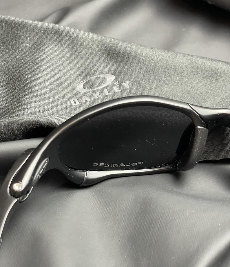 May include: Black sunglasses with a curved frame and dark lenses. The word "POLARIZED" is printed on the lens. A black Oakley carrying case is in the background.