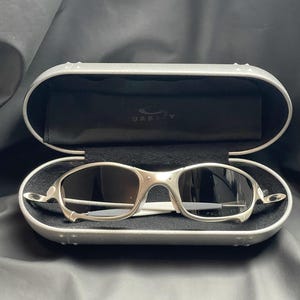 May include: Silver-framed sunglasses with dark lenses rest inside an open, silver case. The case has a black interior with the brand name "Oakley" visible. The sunglasses have a sleek, modern design.