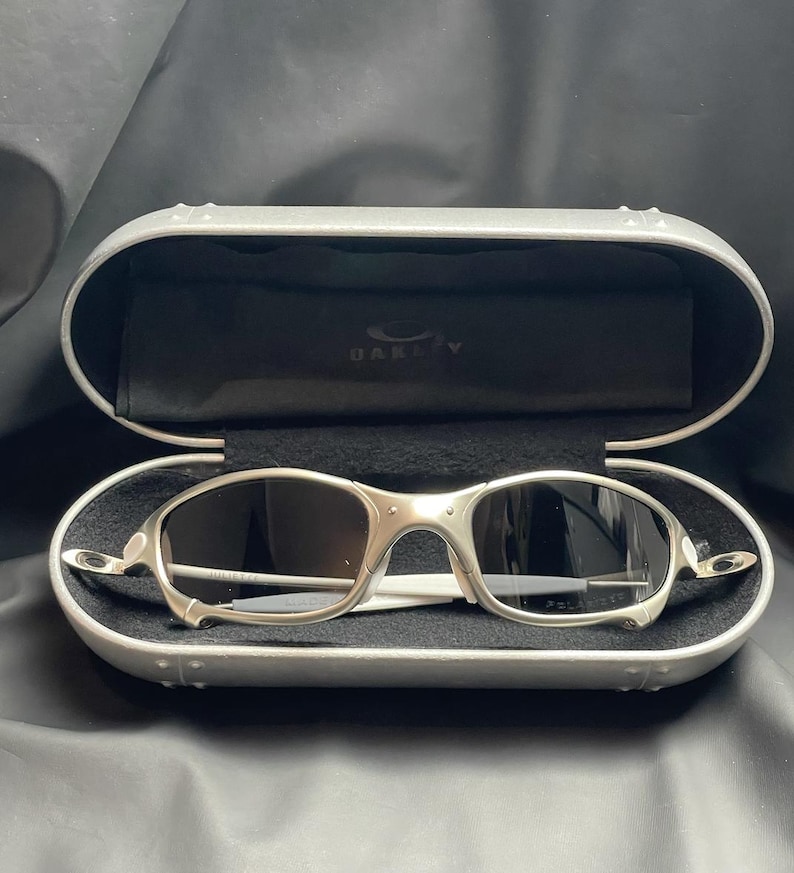 May include: Silver-framed sunglasses with dark lenses rest inside an open, silver case. The case has a black interior with the brand name "Oakley" visible. The sunglasses have a sleek, modern design.