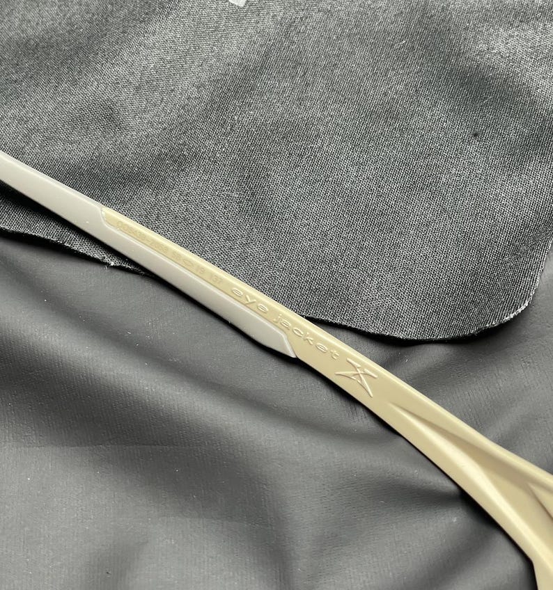 Puede incluir: Primer plano de unas gafas de sol beige y blancas. El texto "eye jacket" est&aacute; impreso en la patilla. Las gafas est&aacute;n sobre una tela gris oscura.