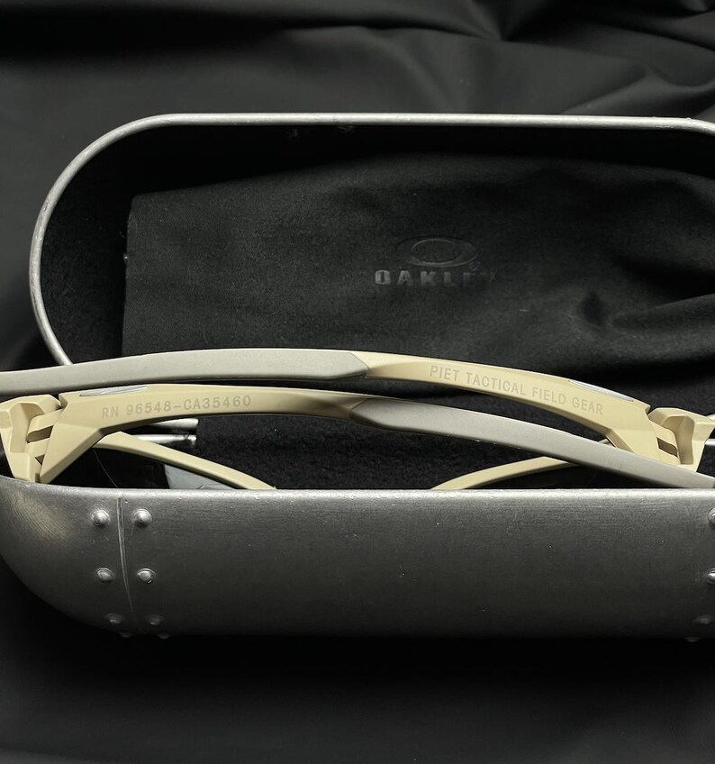 Puede incluir: Gafas de sol beige y grises con el texto "PIET TACTICAL FIELD GEAR" y "RN 96548-CA35460" impreso en la montura. Las gafas de sol est&aacute;n dentro de un estuche plateado con interior negro y un pa&ntilde;o negro.