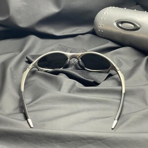 May include: Silver-framed sunglasses with dark lenses, resting on a black cloth. The sunglasses have a sleek, modern design with curved temples. A matching silver case is visible in the background, suggesting a protective storage solution for the eyewear.