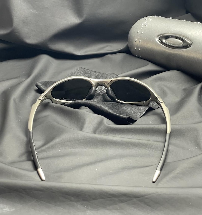 May include: Silver-framed sunglasses with dark lenses, resting on a black cloth. The sunglasses have a sleek, modern design with curved temples. A matching silver case is visible in the background, suggesting a protective storage solution for the eyewear.