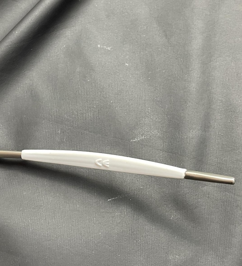 May include: A close-up of a medical instrument with a white handle and dark gray metal ends. The handle has the letters "CE" printed on it. The instrument is on a dark gray surface.