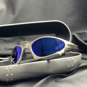 May include: Silver-framed sunglasses with blue lenses rest in an open, gray case. The sunglasses have a sleek, modern design. The case is lined with black fabric. The text "MADE IN USA" is visible on the frame.