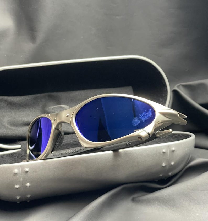 May include: Silver-framed sunglasses with blue lenses rest in an open, gray case. The sunglasses have a sleek, modern design. The case is lined with black fabric. The text "MADE IN USA" is visible on the frame.