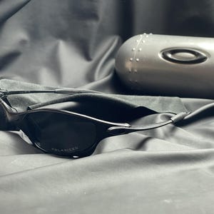 May include: Black sunglasses with a polarized lens and a sleek, modern design. The sunglasses are accompanied by a silver case with the Oakley logo. The case is oval-shaped and has a textured surface.