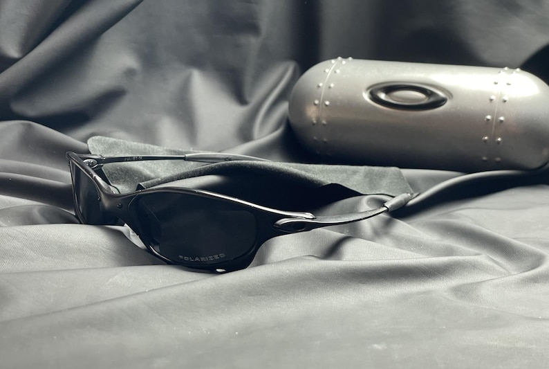 May include: Black sunglasses with a polarized lens and a sleek, modern design. The sunglasses are accompanied by a silver case with the Oakley logo. The case is oval-shaped and has a textured surface.