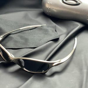 May include: Silver-framed sunglasses with dark lenses rest on a black cloth, next to a silver case. The sunglasses have a sleek, modern design. The case is oval-shaped and has a metallic finish.