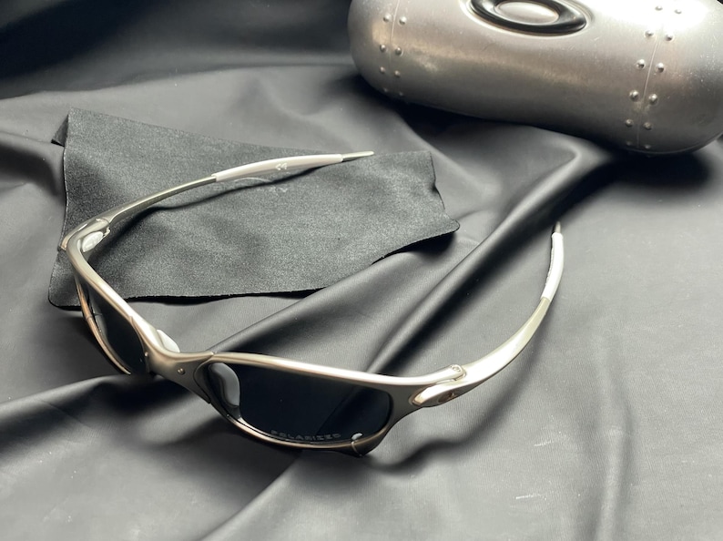 May include: Silver-framed sunglasses with dark lenses rest on a black cloth, next to a silver case. The sunglasses have a sleek, modern design. The case is oval-shaped and has a metallic finish.