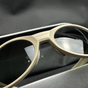 Puede incluir: Primer plano de gafas de sol beige con lentes oscuras. Las gafas de sol tienen un dise&ntilde;o moderno con una montura gruesa y la palabra "PET" impresa en el puente. Las lentes son reflectantes.