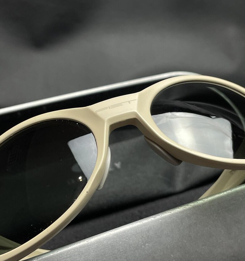 Puede incluir: Primer plano de gafas de sol beige con lentes oscuras. Las gafas de sol tienen un dise&ntilde;o moderno con una montura gruesa y la palabra "PET" impresa en el puente. Las lentes son reflectantes.