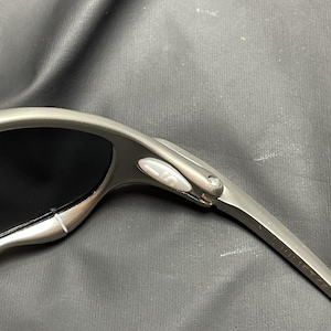 May include: Close-up of a pair of silver sunglasses with black lenses. The sunglasses have a sleek, modern design with a curved frame and a small, white button detail near the hinge.