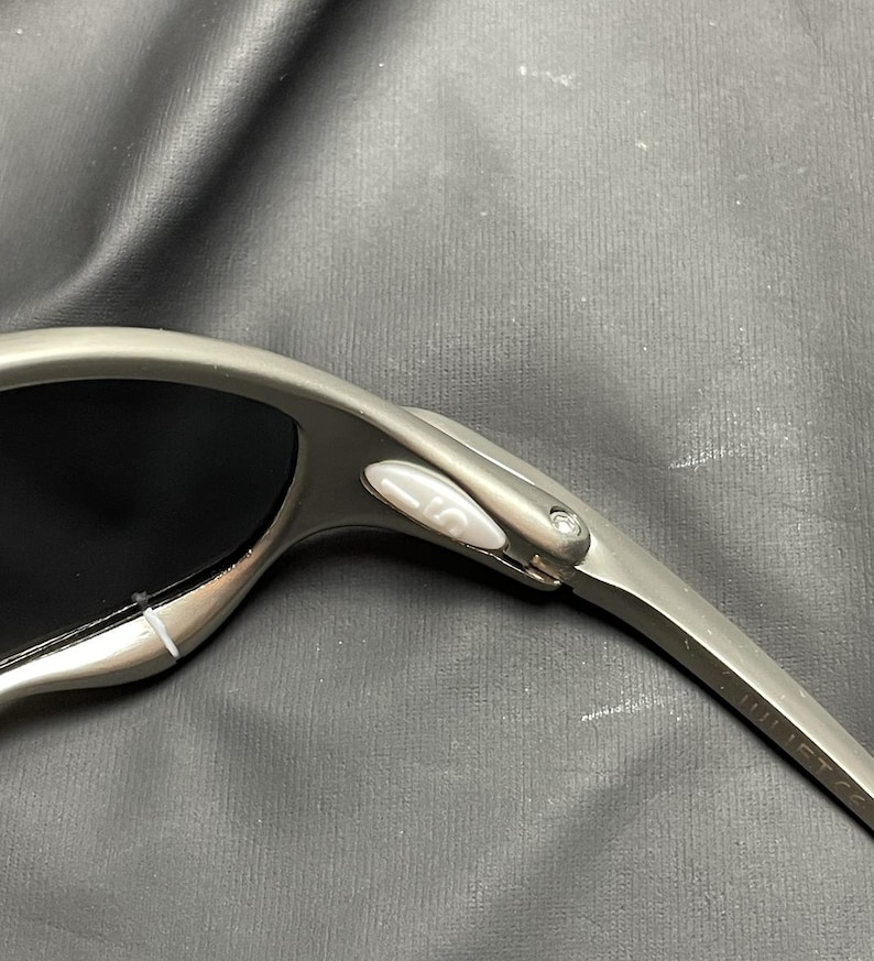 May include: Close-up of a pair of silver sunglasses with black lenses. The sunglasses have a sleek, modern design with a curved frame and a small, white button detail near the hinge.
