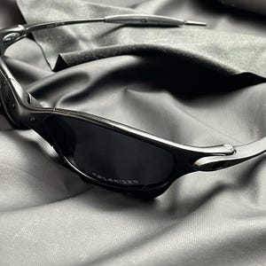 May include: Black sunglasses with polarized lenses. The sunglasses have a sleek, modern design with a dark frame and lenses. The word "POLARIZED" is printed on the lens. The sunglasses are resting on a dark gray cloth.