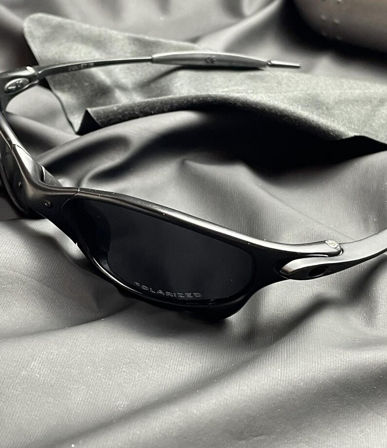 May include: Black sunglasses with polarized lenses. The sunglasses have a sleek, modern design with a dark frame and lenses. The word "POLARIZED" is printed on the lens. The sunglasses are resting on a dark gray cloth.