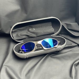 May include: Silver-framed sunglasses with blue lenses rest inside an open, silver case. The case is lined with black fabric and a black cleaning cloth. The Oakley logo is visible on the cloth. The background is a black, textured surface.