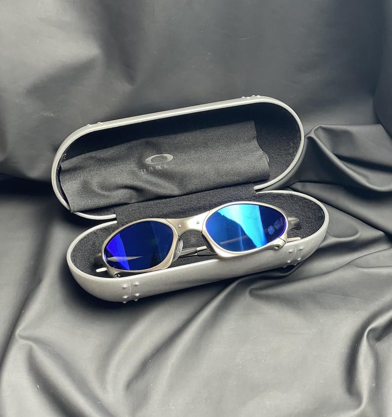 May include: Silver-framed sunglasses with blue lenses rest inside an open, silver case. The case is lined with black fabric and a black cleaning cloth. The Oakley logo is visible on the cloth. The background is a black, textured surface.