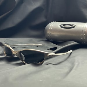 May include: Silver-framed sunglasses with dark polarized lenses and a matching silver case. The sunglasses have a sleek, modern design. The case is metallic with a riveted design and a black oval logo.