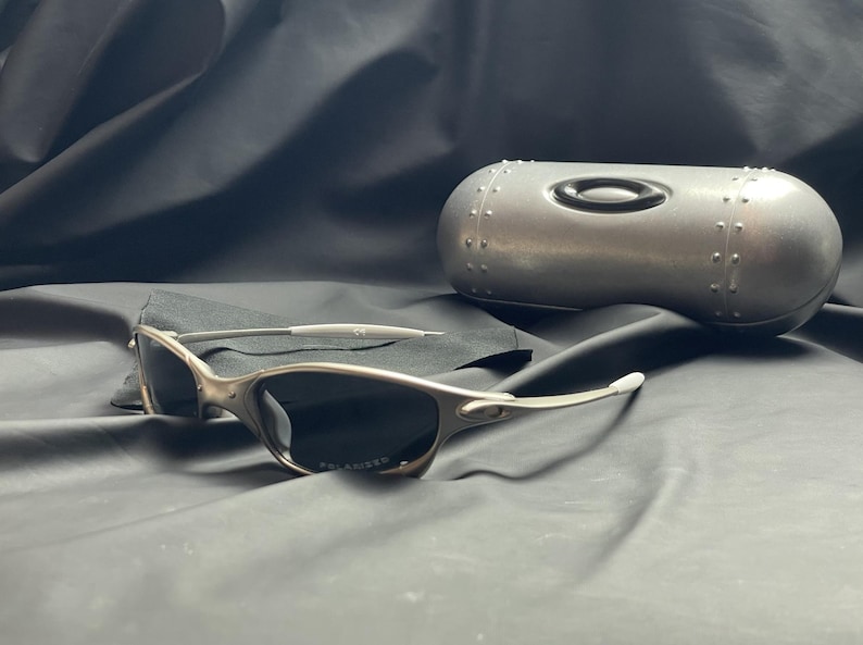 May include: Silver-framed sunglasses with dark polarized lenses and a matching silver case. The sunglasses have a sleek, modern design. The case is metallic with a riveted design and a black oval logo.