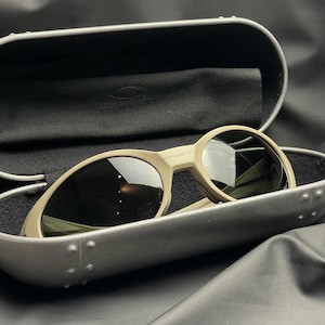 Puede incluir: Gafas de sol color beige con lentes oscuras dentro de un estuche gris. Las gafas de sol tienen un dise&ntilde;o de montura redondeada. El estuche est&aacute; abierto, revelando un interior negro y un pa&ntilde;o suave. La marca es visible en las gafas.