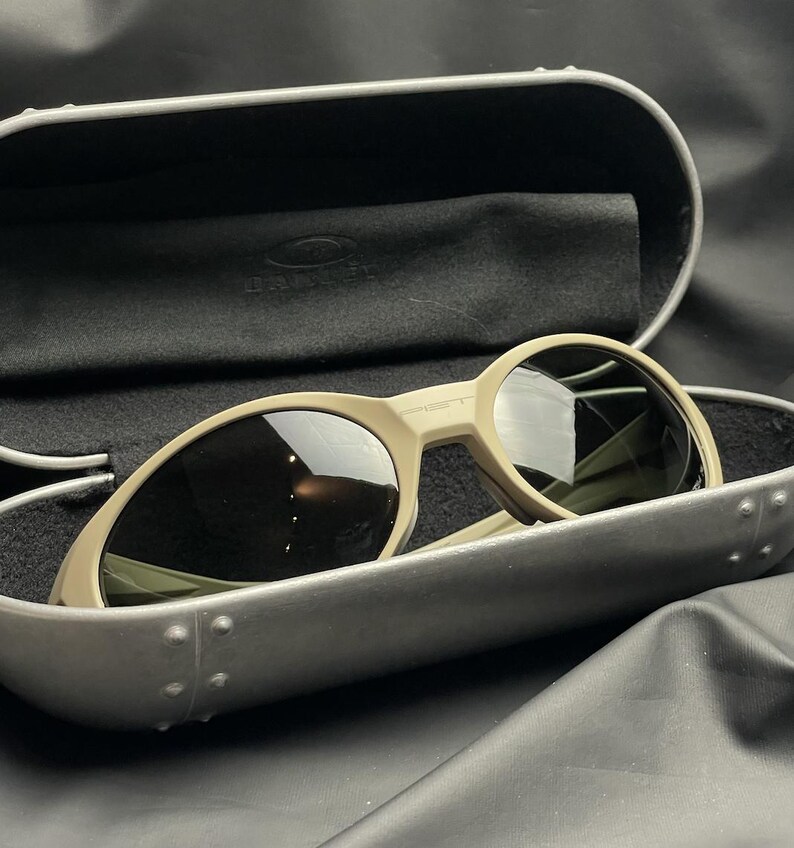 Puede incluir: Gafas de sol color beige con lentes oscuras dentro de un estuche gris. Las gafas de sol tienen un dise&ntilde;o de montura redondeada. El estuche est&aacute; abierto, revelando un interior negro y un pa&ntilde;o suave. La marca es visible en las gafas.