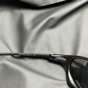 May include: Black sunglasses with a polarized lens. The frame is black and has the numbers "04-114A 56-21 135" printed on the arm. The sunglasses are set against a dark gray background.