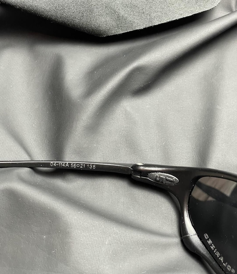 May include: Black sunglasses with a polarized lens. The frame is black and has the numbers "04-114A 56-21 135" printed on the arm. The sunglasses are set against a dark gray background.
