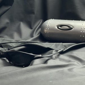 May include: Black sunglasses with a polarized lens, resting on a dark cloth surface. A silver, cylindrical case with the Oakley logo and rivet detailing is also present. The overall aesthetic is sleek and modern.