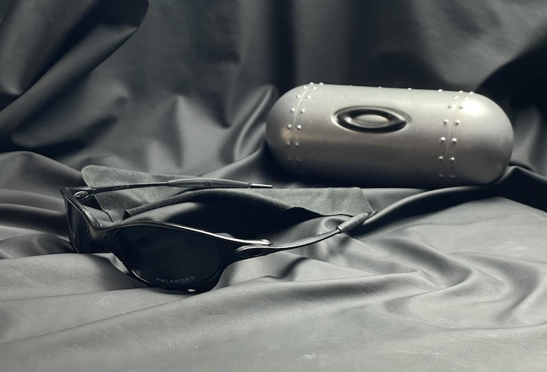 May include: Black sunglasses with a polarized lens, resting on a dark cloth surface. A silver, cylindrical case with the Oakley logo and rivet detailing is also present. The overall aesthetic is sleek and modern.