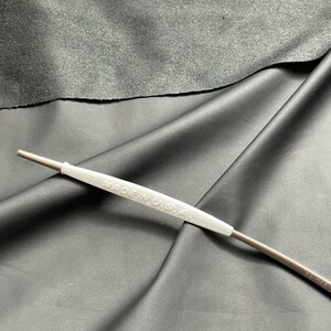 May include: A metal tool with a white handle that reads "MADE IN U.S.A." is displayed against a dark gray background. The tool has a thin, cylindrical body and a pointed tip. The handle is a light gray color.