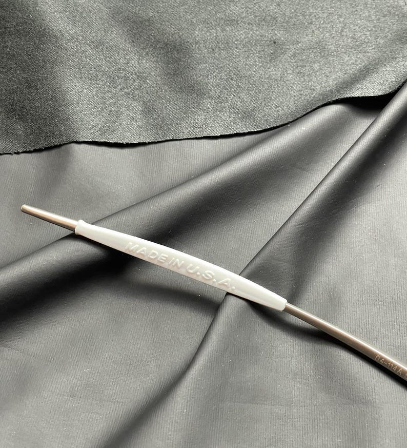 May include: A metal tool with a white handle that reads "MADE IN U.S.A." is displayed against a dark gray background. The tool has a thin, cylindrical body and a pointed tip. The handle is a light gray color.