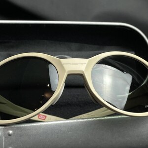 Puede incluir: Gafas de sol de color beige con lentes oscuras. Las gafas tienen una montura redondeada y la palabra "PET" est&aacute; impresa en el puente. La montura tiene una marca de EE. UU. en la patilla izquierda. Las gafas est&aacute;n en un estuche plateado.