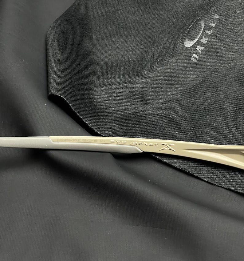 Puede incluir: Primer plano de unas gafas de sol Oakley con montura gris claro. El texto "eye jacket" es visible en la patilla. Una bolsa de microfibra negra Oakley est&aacute; en el fondo.