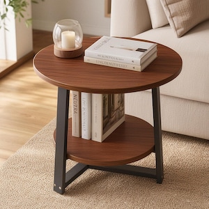 Puede incluir: Una mesa auxiliar redonda con tablero y estante inferior de madera oscura, sostenida por patas de metal negro. En la superficie superior hay un portavelas de cristal con una vela blanca y dos libros titulados Kinfolk y Living Beautifully.