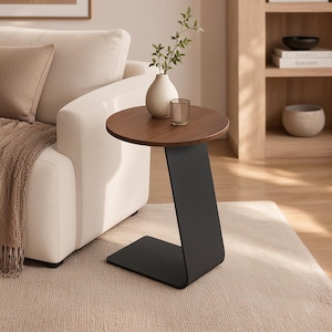 May include: A C-shaped side table featuring a circular dark brown wooden top supported by a single matte black metal base that curves to form a flat rectangular floor stand. The table surface holds a small beige ceramic vase and a glass tumbler.