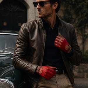 Könnte beinhalten: Braune Lederjacke mit Stehkragen und rote Fahrerhandschuhe mit braunen Akzenten. Die Handschuhe haben kreisförmige Perforationen. Die Person trägt eine Sonnenbrille und ein schwarzes Poloshirt. Khakihose und ein Oldtimer sind ebenfalls zu sehen.