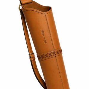 May include: A tan leather quiver featuring a cylindrical body, a stamped arrow icon, and a decorative cross-stitched band. The quiver includes an adjustable shoulder strap with a brass buckle and two brass rivets at the top attachment point.