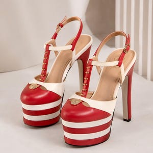 May include: A pair of red and white platform high-heeled sandals featuring a T-strap design, an adjustable slingback strap with a gold buckle, and a gold-toned lion head emblem centered on the rounded toe box of each shoe.