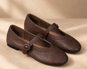 May include: A pair of dark brown flat shoes featuring a rounded toe and a single strap across the instep. The strap is secured with a matching circular buckle. The shoes have a smooth, matte finish and a low-profile design with a thin black sole.