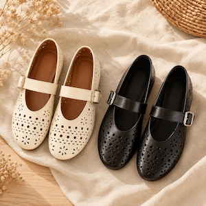 May include: Two pairs of Mary Jane shoes, one in cream and one in black. Each shoe features a rounded toe, a single strap with a gold-toned metal buckle, and a laser-cut floral and circular pattern across the front and sides of the upper.