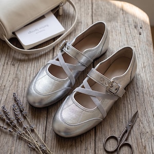 May include: A pair of metallic silver flats featuring a rounded toe, a buckled strap across the instep, and crisscrossing elastic bands. The shoes have a textured finish and a low profile, providing a sleek look for various occasions.