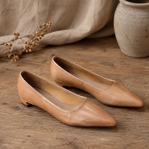 Op de afbeelding: Een paar platte schoenen van beige leer met een puntige neus en een lage blokhak. De schoenen hebben een gladde, matte afwerking met zichtbare stiksels langs de bovenrand en een gestructureerd silhouet met een eenvoudig ontwerp.