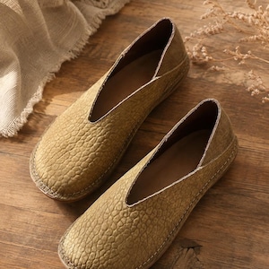 Puede incluir: Un par de zapatos sin cordones de color verde oliva con una superficie texturizada similar al cuero granulado, un corte profundo en forma de V en el empeine y costuras claras visibles a lo largo de la punta redondeada y el borde de la suela.