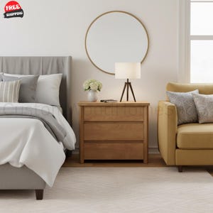 May include: A light brown wooden nightstand with three drawers, positioned between a gray upholstered bed and a yellow armchair. A white lamp with a tripod base sits on top, next to a white vase with flowers, beneath a round wall mirror.
